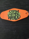 Stay Weird bold orange motel-style keychain with sage green starburst background and chunky black hand-lettered text