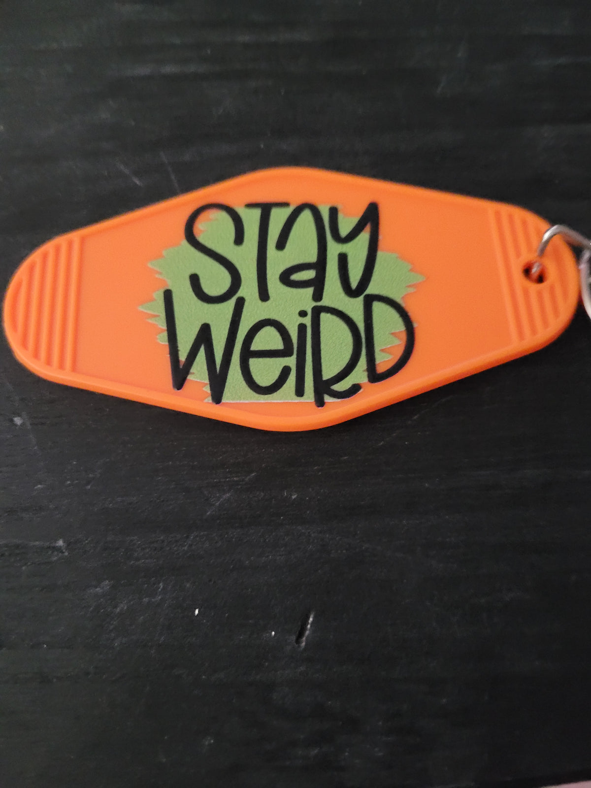 Stay Weird bold orange motel-style keychain with sage green starburst background and chunky black hand-lettered text